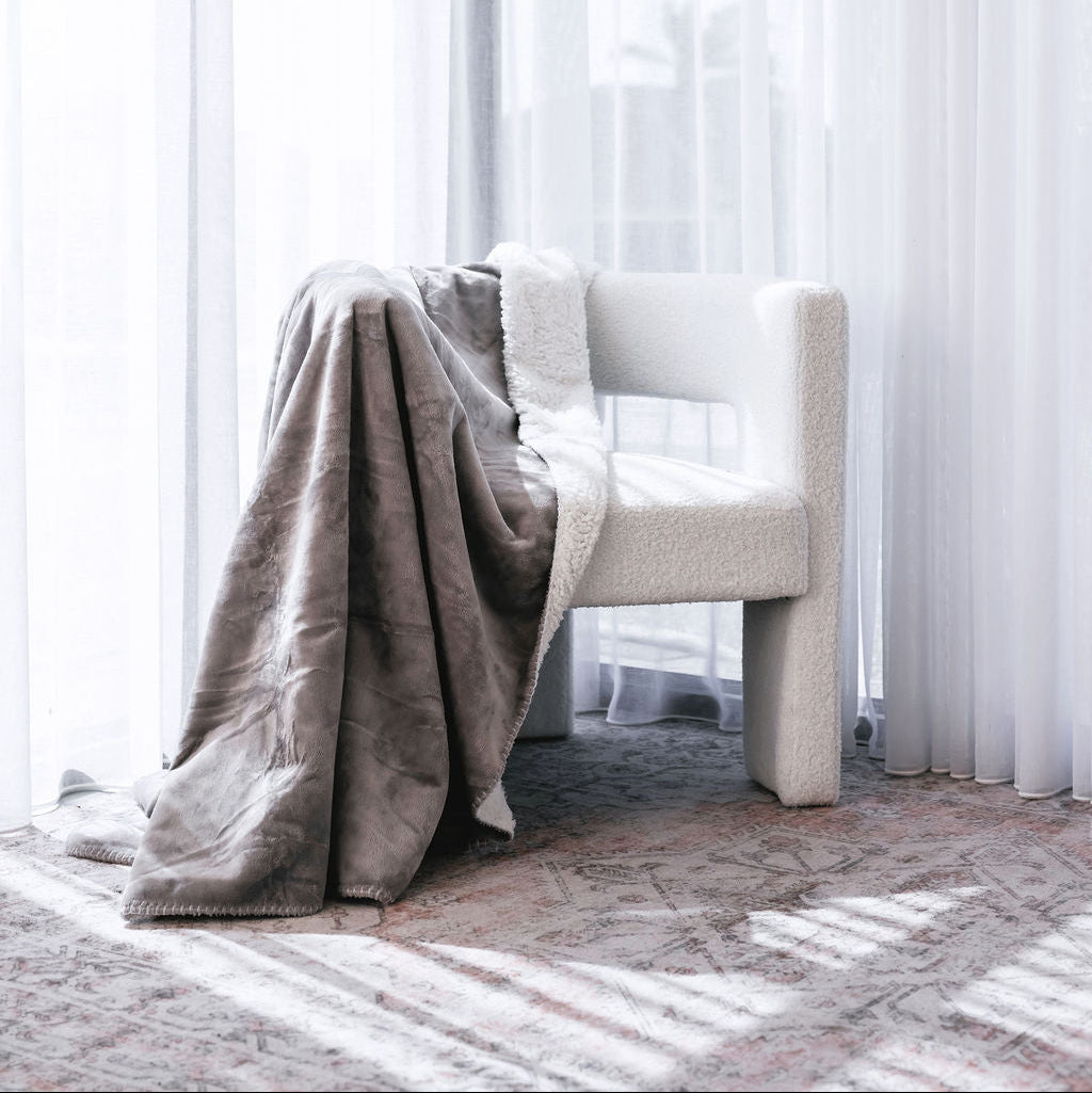 A soft taupe-gray throw blanket draped over a cream-colored armchair in a bright, sunlit room with sheer white curtains and a patterned rug.