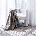 A soft taupe-gray throw blanket draped over a cream-colored armchair in a bright, sunlit room with sheer white curtains and a patterned rug.